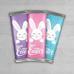 Puede incluir: Tres envoltorios de barras de chocolate con temática de Pascua en morado, rosa y azul. Cada envoltorio presenta un conejito blanco sonriente y las palabras "Happy Easter" en una fuente cursiva. Los envoltorios tienen una parte superior e inferior de papel de aluminio plateado.