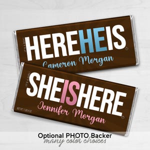 May include: Two chocolate bars with text that reads "HEREHEIS" and "SHEISHERE". The first bar has blue text and the second has pink text. Both bars have a name printed below the main text. The bars are wrapped in silver foil.