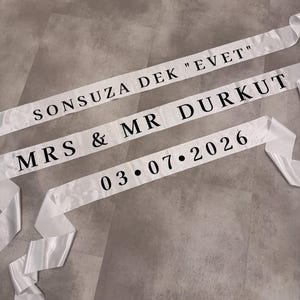 Könnte beinhalten: Drei weiße Satinschärpen mit schwarzem Textaufdruck. Die oberste Schärpe trägt die Aufschrift SONSUZA DEK "EVET", die mittlere MRS & MR DURKUT und die untere zeigt das Datum 03.07.2026 in einer fetten, serifenlosen Schriftart.