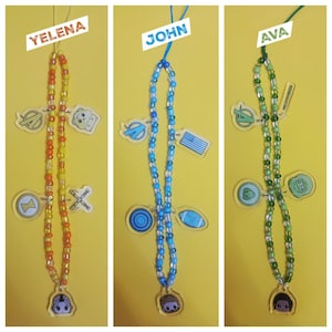 Könnte beinhalten: Drei Perlen-Lanyards mit Charakteranhängern und Namensschildern. Ein Lanyard ist orange und gelb, eines ist blau und eines ist grün. Jedes Lanyard hat einen anderen Charakteranhänger und ein Namensschild mit der Aufschrift "YELENA", "JOHN" und "AVA".