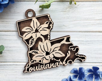 May include: Wooden ornament shaped like the state of Louisiana with a floral design and the word "Louisiana" written below. The ornament is on a white wooden background.
