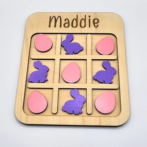 May include: A wooden tic-tac-toe game with the name Maddie engraved on the top. The game board has nine spaces with purple bunnies and pink eggs as the game pieces.