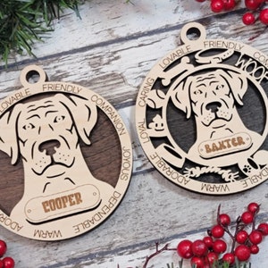 May include: Two wooden dog ornaments with a laser-cut design. The ornaments are round and feature a dog's head with the words "Loyal, Caring, Adorable, Friendly, Companion, Joyous, Dependable, Warm" around the edge. The ornaments are personalized with the names "Cooper" and "Baxter".