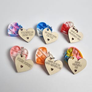 May include: Six heart-shaped pop-it fidget toys with wooden heart tags. The tags have different Valentine's Day messages, including "You Make My Heart Pop!" and "Have a Poppin' Valentine's Day!"