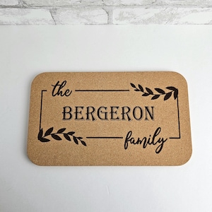 May include: A cork doormat with the text "the Bergeron family" in a black frame with leafy accents.
