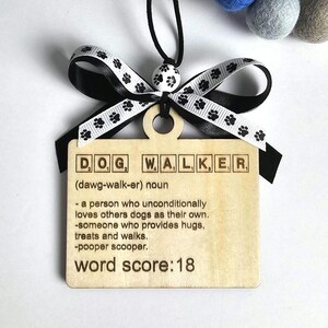 May include: Wooden plaque with the text "DOG WALKER" in Scrabble-style tiles. The plaque includes a definition and a word score of 18. It is adorned with a black and white paw print ribbon and a black cord for hanging.