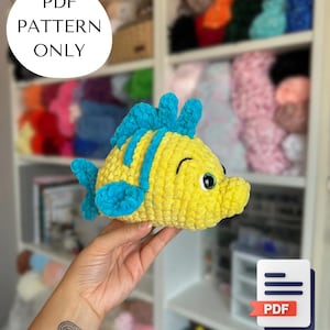 Puede incluir: Un pez amarillo tejido a ganchillo con aletas y detalles azules. El pez se sostiene en una mano. La imagen incluye el texto "PDF PATTERN ONLY" en un círculo blanco y un icono PDF con una pancarta roja.
