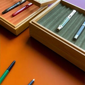Peut inclure: Des présentoirs à stylos en bois avec couvercles en verre, présentant divers stylos plume. Les étuis ont des intérieurs en velours orange et vert. Plusieurs stylos sont visibles, dont des stylos noirs, rouges et argentés, avec quelques stylos posés sur la surface orange.