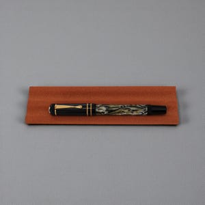 May include: A fountain pen featuring a black cap with a gold-coloured clip and trim, paired with a barrel displaying a marbled pattern of black, grey, and white tones. The pen rests horizontally on a rectangular, textured, rust-coloured tray.