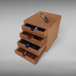 May include: A wooden desk organizer with four tiered drawers, each featuring a blue interior lining. The unit is crafted from light brown wood with a visible grain. A black and silver fountain pen rests on the top surface of the organizer.