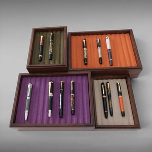 May include: Four dark wooden display trays with corrugated velvet inserts in olive green, orange, purple, and beige. Each tray holds a collection of fountain pens with various barrel designs, metallic clips, and caps in black, silver, and patterned finishes.