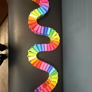 May include: A long, serpentine paper chain decoration featuring a vibrant rainbow gradient. The chain is constructed from individual interlocking paper loops in shades of red, orange, yellow, green, blue, and purple, arranged in a repeating pattern.