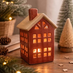 Op de afbeelding: Een roodkleurige keramische kandelaar in de vorm van een huis, met een beige dak en schoorsteen. Het huis heeft meerdere vierkante ramen die een warme gloed uitstralen. Decoratieve dennenbomen en dennenappels zijn op de achtergrond.