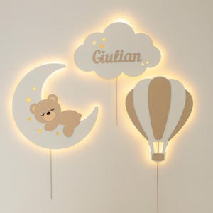 May include: A nursery wall decor set featuring a crescent moon with a sleeping bear, a cloud with the name "Giulian", and a hot air balloon. The set is in neutral colors with warm lighting.