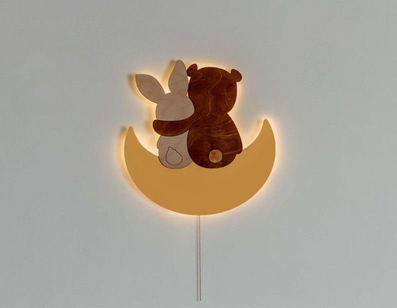 May include: A wooden wall light shaped like a crescent moon with a brown bear and a bunny rabbit sitting on top. The light is illuminated with a warm yellow glow.