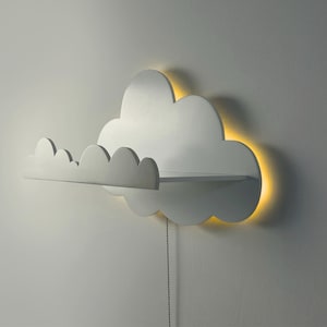 May include: A white cloud-shaped wall shelf with a built-in light. The shelf has a scalloped edge and a small shelf area. The light emits a warm glow around the cloud's edges. A pull chain hangs below.