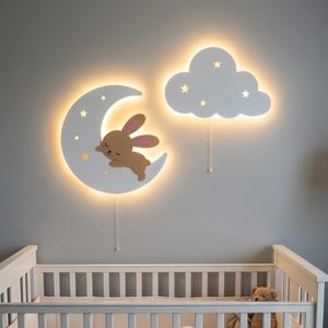 Op de afbeelding: Twee witte LED-wandlampen in de vorm van een halve maan met een slapend bruin konijn en een wolk. Beide armaturen hebben stervormige uitsparingen die een warm licht uitstralen en zijn elk voorzien van een trekkoord aan de onderzijde.