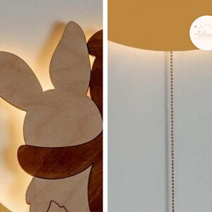 May include: A wooden night light shaped like a bunny and a bear hugging each other. The light is illuminated and casts a warm glow. The night light is mounted on a white wall.