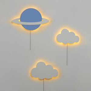 May include: Three wall-mounted light fixtures shaped like a planet and two clouds. The planet is blue with a white ring around it. The clouds are white. All three fixtures have a warm yellow glow.