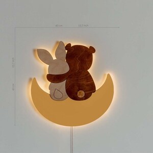 May include: A wooden wall-mounted night light shaped like a crescent moon with a bear and bunny hugging on top. The light is illuminated with a warm yellow glow.