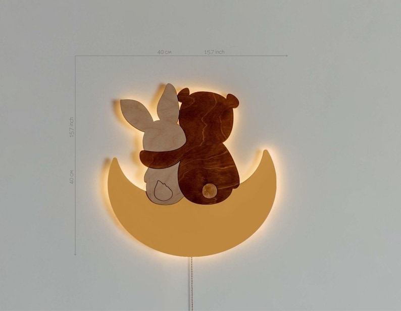 May include: A wooden wall-mounted night light shaped like a crescent moon with a bear and bunny hugging on top. The light is illuminated with a warm yellow glow.