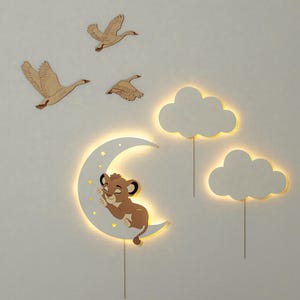 Pode incluir: Um conjunto de decoração de parede de madeira branca com uma lua crescente, nuvens e gansos. A lua tem um filhote de leão dormindo sobre ela. As nuvens e a lua têm luzes brancas quentes.