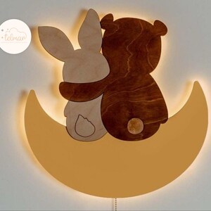 May include: A wooden night light shaped like a crescent moon with a bear and a rabbit sitting on top. The bear and rabbit are hugging and the moon is lit up with a warm glow.