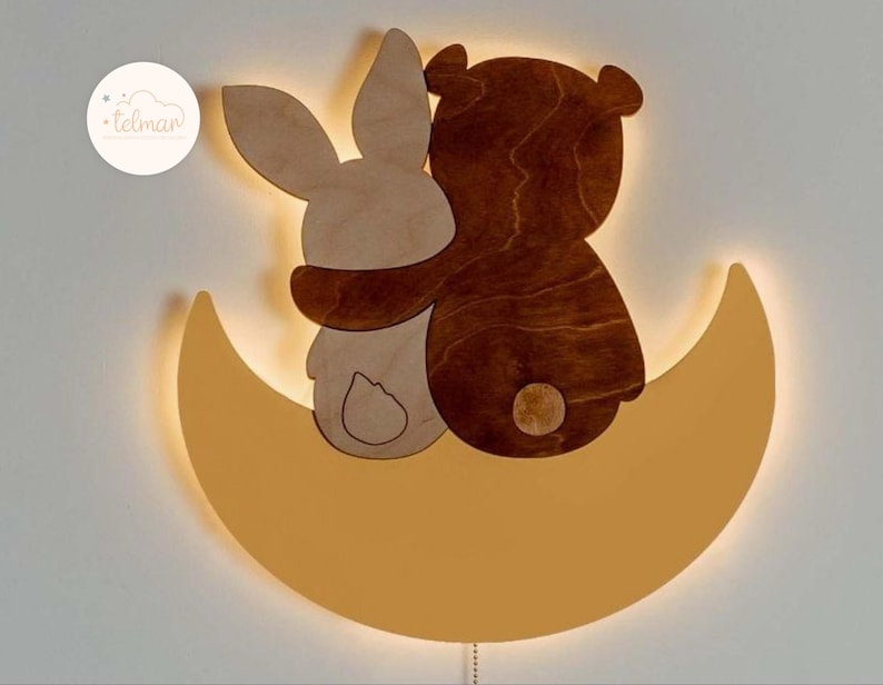 May include: A wooden night light shaped like a crescent moon with a bear and a rabbit sitting on top. The bear and rabbit are hugging and the moon is lit up with a warm glow.