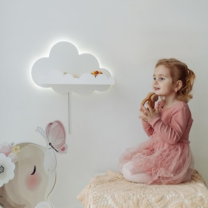 Pode incluir: Uma criança com um vestido rosa está sentada em um cobertor bege, segurando um brinquedo de madeira. Uma prateleira branca em forma de nuvem com luz está montada na parede. Uma cabeça de veado decorativa com uma borboleta também é visível.