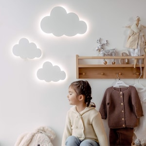 Puede incluir: Una niña pequeña está sentada frente a una pared blanca con tres luces blancas en forma de nube. Encima de ella hay una estantería de madera con ganchos, con una muñeca y otras decoraciones. Dos cárdigans de punto cuelgan de los ganchos.