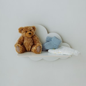 Pode incluir: Uma prateleira branca em forma de nuvem com um urso de peluche castanho sentado nela. Há sapatinhos de bebé de malha azul e uma fralda de pano branca na prateleira.