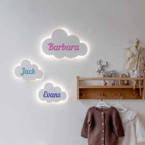 Könnte beinhalten: Drei weiße, wolkenförmige Wandleuchten mit den Namen "Barbara", "Jack" und "Evans" in bunten Buchstaben. Die Leuchten sind beleuchtet und an einer weißen Wand montiert. Ein Holzregal mit Haken ist unterhalb der Leuchten an der Wand montiert.