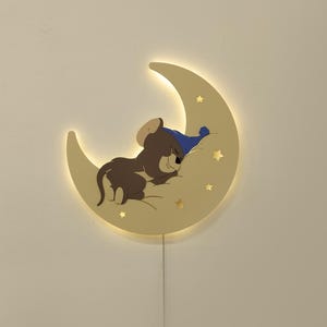 以下が含まれることがあります： 三日月型の壁掛け照明。眠っている漫画のキャラクターが特徴です。光は温かみのある黄色で、キャラクターは茶色で青いナイトキャップをかぶっています。月には星の切り抜きがいくつかあります。