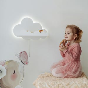 Pode incluir: Uma luz noturna branca em forma de nuvem com uma prateleira, montada em uma parede branca. Uma menina está sentada em um tapete branco, segurando um brinquedo de madeira. Um elefante rosa e marrom com um adesivo de borboleta está na parede.