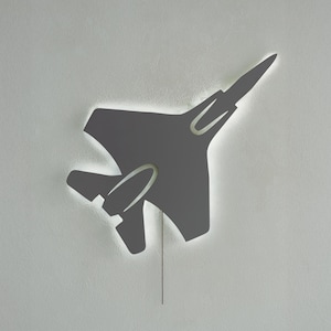 Pode incluir: Decoração de parede cinza em forma de avião com um contorno iluminado. A silhueta do jato é visível, com uma corrente pendurada por baixo. A decoração é montada numa parede cinza claro.