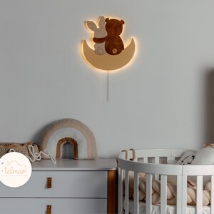 May include: A wooden wall-mounted night light shaped like a crescent moon with a bear and bunny cuddling inside. The light is illuminated and casts a warm glow.