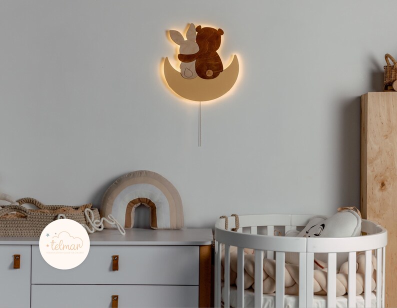 May include: A wooden wall-mounted night light shaped like a crescent moon with a bear and bunny cuddling inside. The light is illuminated and casts a warm glow.