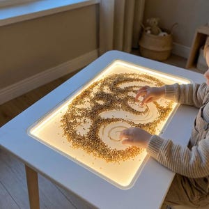 May include: A white, rectangular light table with wooden legs. The table's surface is illuminated, displaying a pattern created with small, tan-colored beads. A child's hands are visible, interacting with the beads.