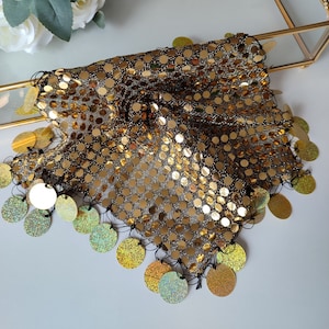 May include: A gold sequined fabric with a fringe of gold sequins around the edges. The fabric is folded in half and has a black thread running through it.