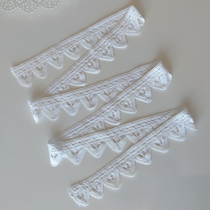 May include: Several strips of white lace with a scalloped edge are arranged diagonally on a white surface. The lace has a delicate, openwork design with heart-shaped details. The lace is likely used for sewing, crafting, or adding a decorative touch to garments.