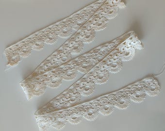 May include: Off-white crocheted lace trim in various lengths. The trim features a scalloped edge and a delicate, openwork design. Perfect for sewing, crafting, or adding a vintage touch to projects. The lace is laid out on a white surface.