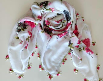 Pode incluir: Um lenço quadrado branco com um padrão floral de rosas cor-de-rosa e cinzento-escuro e folhas verdes. As extremidades do lenço são rematadas com delicadas franjas de missangas pendentes em tons de rosa, branco e verde.