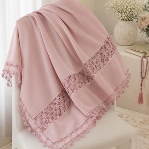May include: A soft, pink shawl with intricate lace detailing along the edges is draped over a white chair. The shawl's delicate fabric is gathered, showcasing its texture and design. The lace trim adds a touch of elegance to the overall aesthetic.