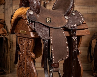 May include: A dark brown leather western saddle with intricate floral tooling, a horn, and a sheepskin underside. The saddle features a metal badge with the text AD and sits on a carved wooden stand with a brass plate reading AD Saddlery-Est 1978.