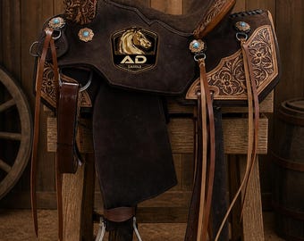 May include: A brown suede western saddle with intricate floral tooling and turquoise accents. The saddle features the "AD SADDLE" logo with a horse head. It is displayed on a wooden stand, showcasing its detailed craftsmanship.