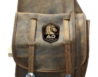 May include: A distressed brown leather saddlebag with a flap closure and buckle details. The bag features a logo patch with a horse head and the text "AD Saddler". The bag has a vintage, worn look.