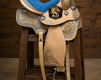 May include: A tan leather saddle with a blue suede seat, detailed floral tooling, and silver-tone embellishments. The saddle has a black emblem with a horse head and the text "AD SADDLE". It is displayed on a dark wooden stool.