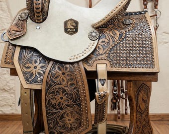 May include: A tan and dark brown leather western saddle featuring intricate floral tooling, silver stud accents, and a cream-colored suede seat. The saddle displays an AD Saddler logo on the seat and horn, with a metal stirrup and leather straps.