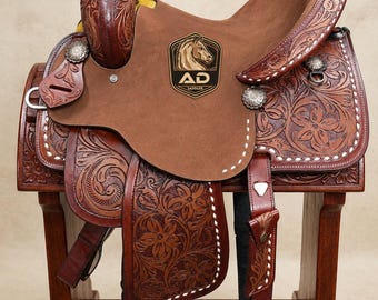 May include: A brown leather western saddle featuring intricate floral tooling, a smooth suede seat, and silver-toned conchos. The saddle displays a black and gold emblem with a horse illustration and the text AD SADDLER on the seat and base.