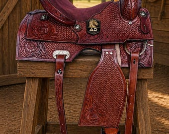 May include: A detailed, reddish-brown leather saddle with intricate floral and geometric patterns. The saddle features a burgundy suede seat and stirrups, and is displayed on a wooden stand. A metal emblem with a horse head is visible.